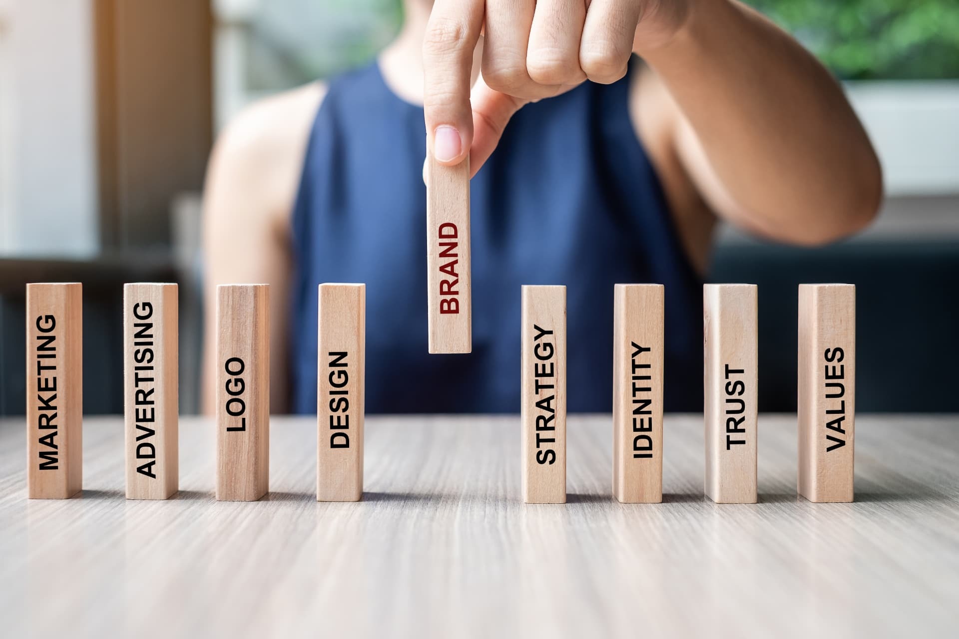 A hand placing a wooden block marked Brand in a row with Marketing, Advertising, Logo, Design, Strategy, Identity, Trust and Values.