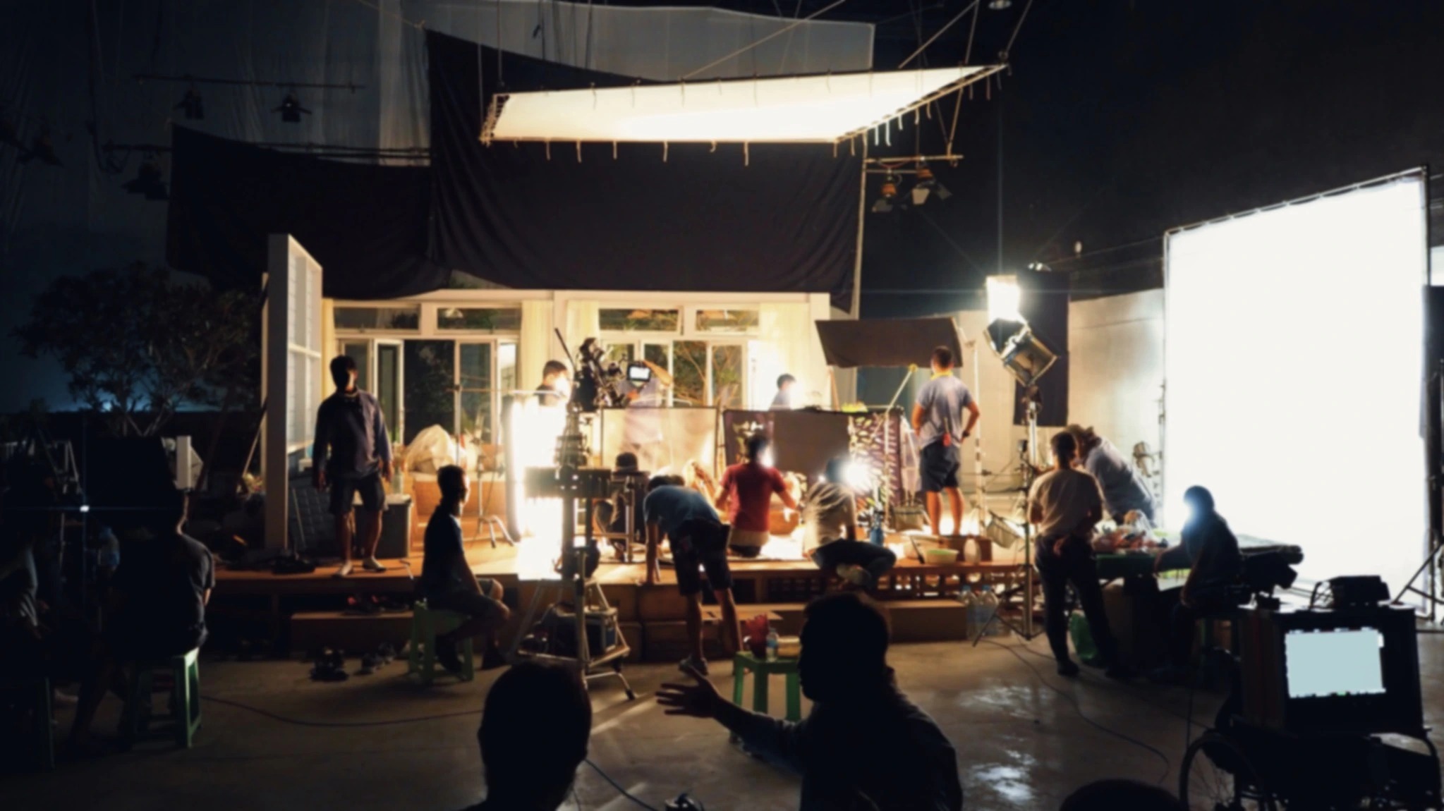 A film crew at work on a lit interior set at night, with large soft lights, cameras and a monitor