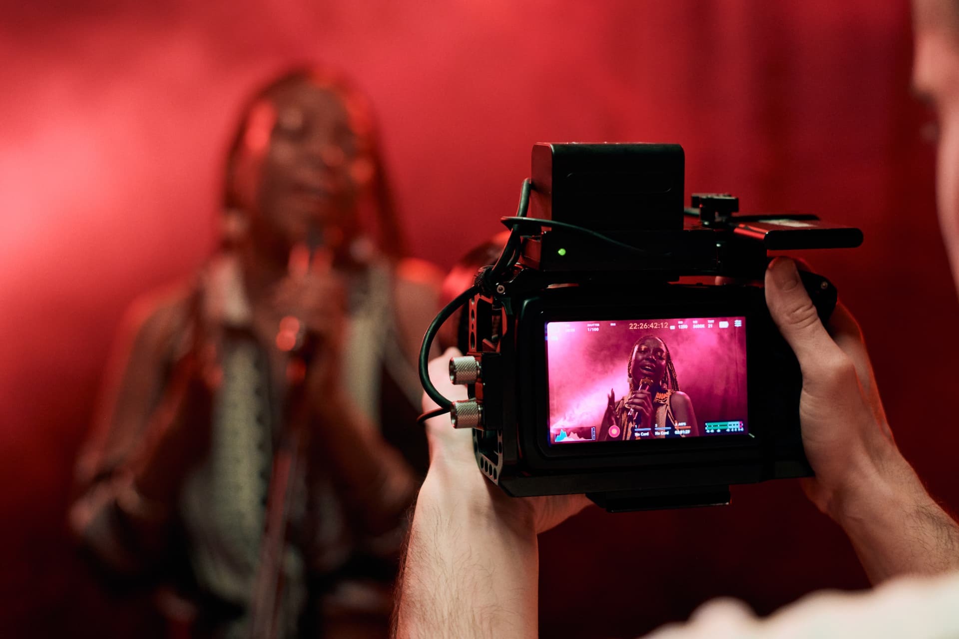 Camera operator filming a singer under red light, the performance shown on the camera's screen.