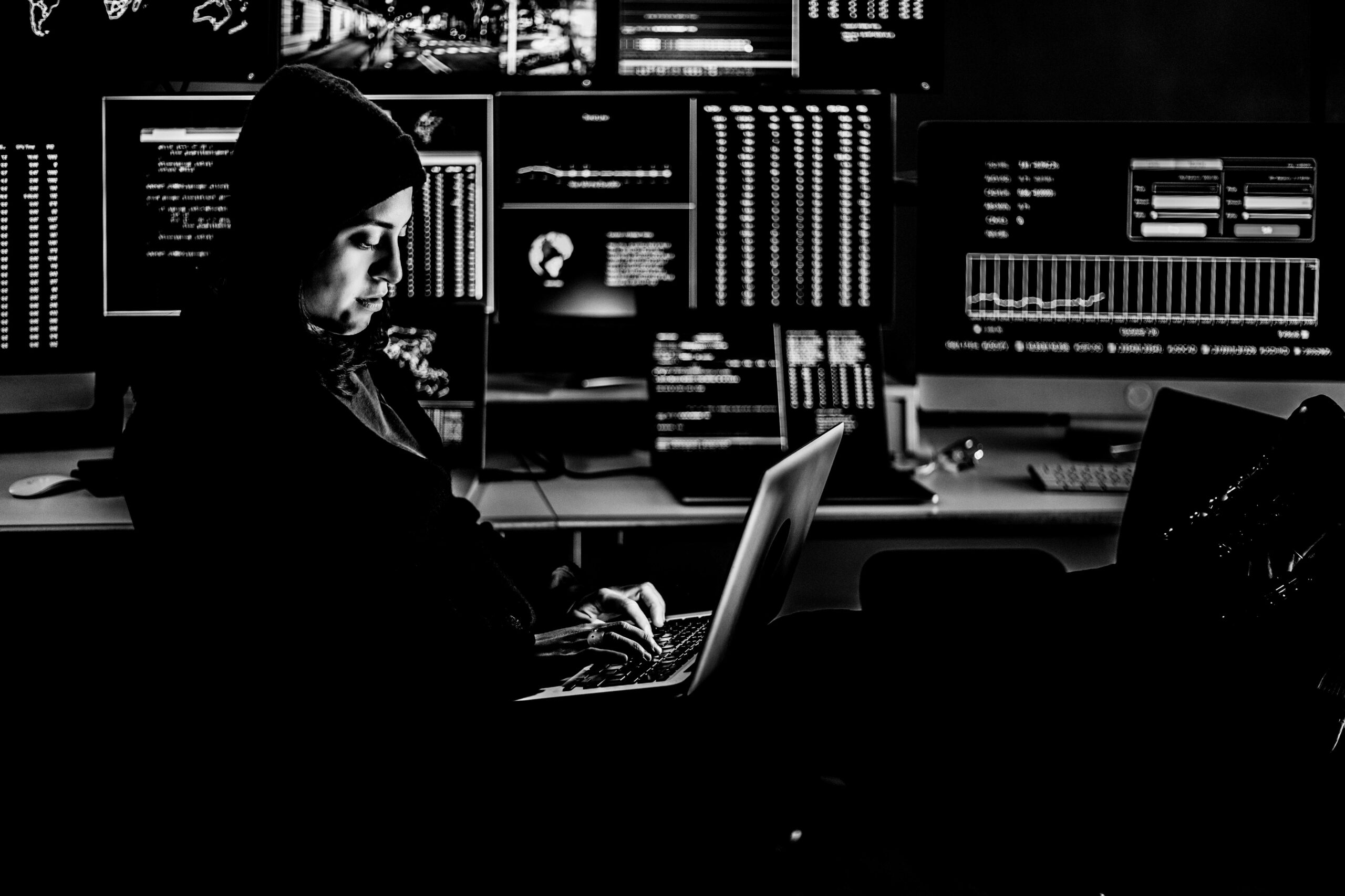 Professional female hacker typing virus code on laptop for cyber attack in dark room with multiple monitors on the background