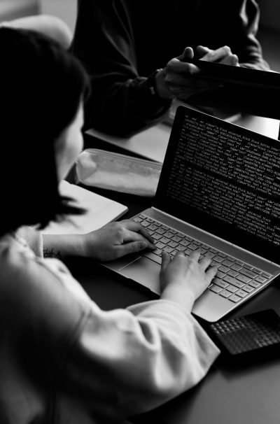 Young female student typing on laptop keyboard while decoding data on screen against two classmates or colleagues discussing presentation